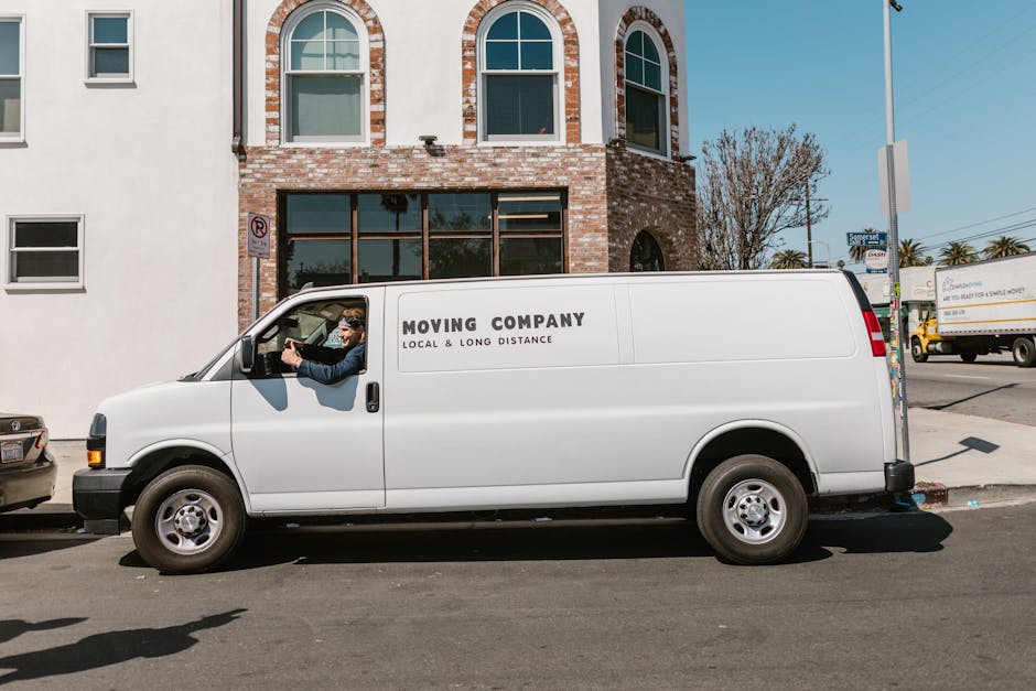 Inside a white moving van operated by Holloway Removals, two professional movers are visible, with one man seated in the driver’s seat and another person in the passenger seat, both smiling at the c
