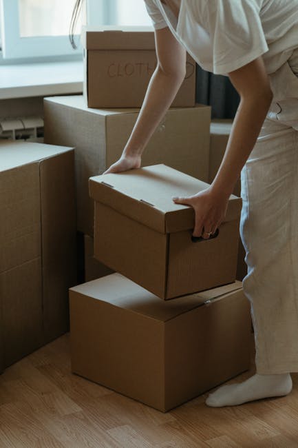 A person wearing light-colored clothing and socks is engaged in a home relocation task, lifting a medium-sized cardboard box with no visible labels or markings from a stack of similar boxes, some with