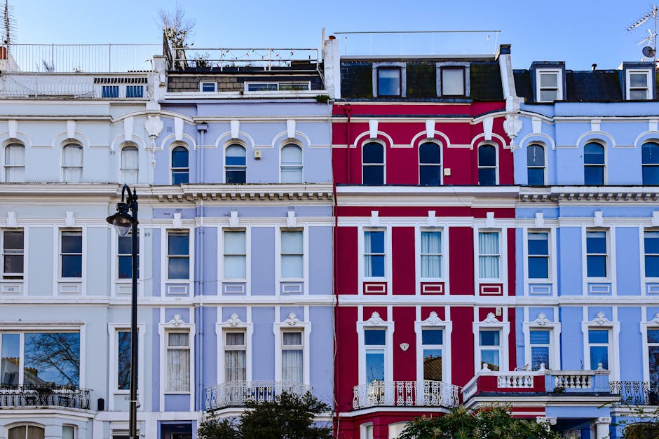 A row of multi-storey Victorian terraced houses with ornate bay windows, painted in various pastel shades including light blue, cream, and bright green, located on a city street during daylight. A bla
