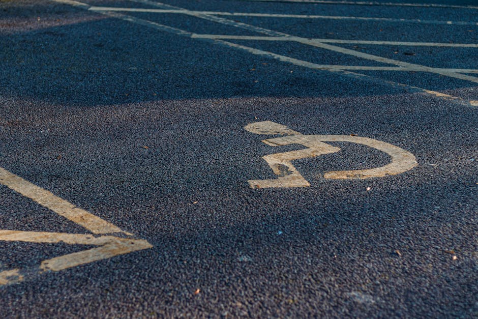 Close-up view of a designated accessible parking space on asphalt pavement, featuring a white wheelchair symbol painted on the surface. The symbol, slightly worn, depicts a person seated in a wheelcha