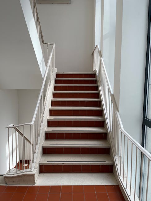 A straight staircase inside a building with white metal railings on both sides, red-tiled steps, and a white wall on the left. Natural light filters through a large window on the right, illuminating t